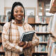 Portrait, black woman and library with book, knowledge and relax with free time. Nigerian female, student smile and customer in bookstore, for reading and literature for education, study and research