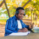 young handsome black male student sitting writing on paper using mobile phone in hand with a colorful school background