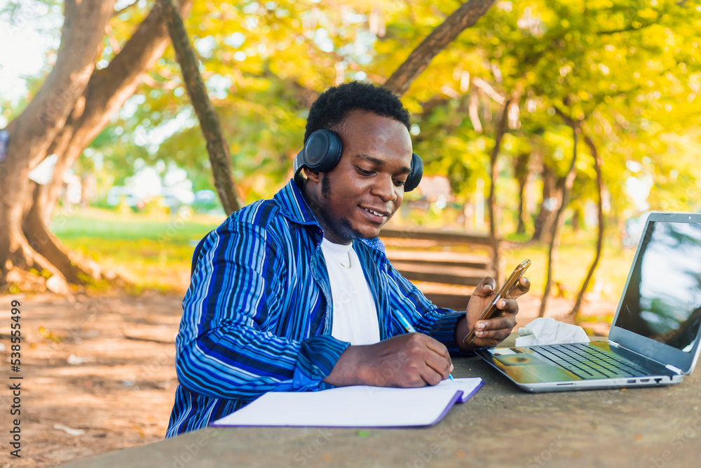 young handsome black male student sitting writing on paper using mobile phone in hand with a colorful school background